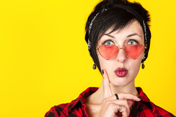 Beautiful portrait of funny brunette woman with heart shaped red glasses holding finger on the chin against yellow background
