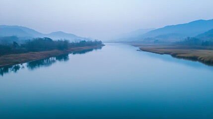 Naklejka premium Calm river flowing through a valley surrounded by misty mountains under a hazy sky in the early morning