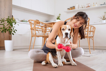 Pretty young woman playing with adorable Beagle dog on yoga mat in kitchen