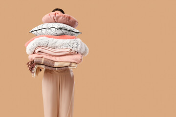 Beautiful young woman with stack of different blankets and pillows on beige background