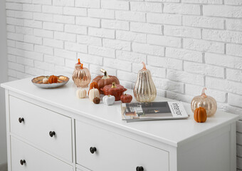 Chest of drawers with candles in shape of pumpkin, decor and magazine near white brick wall