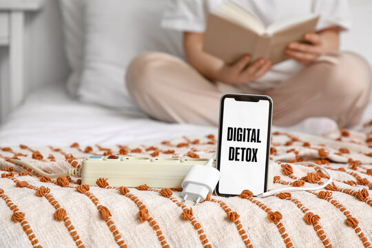 Mobile phone with text DIGITAL DETOX and extension cord against woman reading book in bedroom, closeup. National Day of Unplugging