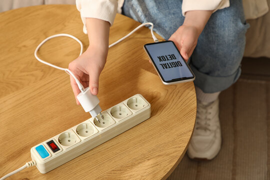 Young woman getting out mobile phone charger from extension cord on table at home, closeup. National Day of Unplugging