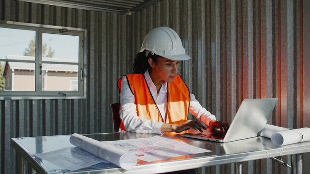 Skilled hispanic woman engineer in safety vest and hard hat working on laptop at modern construction site office
