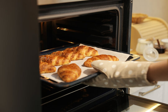 Woman taking baking sheet with tasty croissants out of oven indoors, closeup