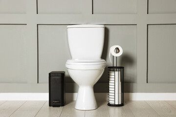 Ceramic toilet bowl, paper and trash can near grey wall indoors