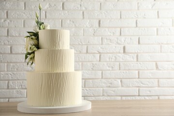 Delicious wedding cake decorated with flowers on wooden table near white brick wall, closeup. Space for text