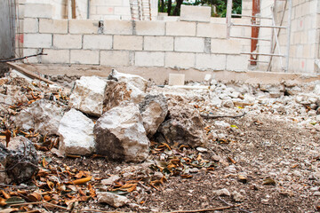 A construction site featuring an incomplete concrete block wall surrounded by raw materials. A pile of large rocks, scattered leaves, and dirt create a rugged foreground, evoking a work-in-progress vi