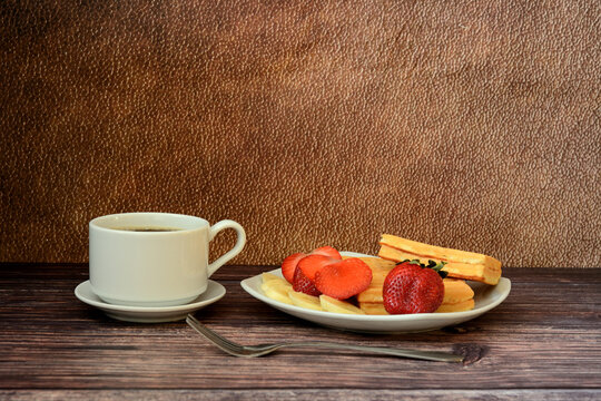 A cup of black coffee on a saucer and a plate with a bunch of waffles with strawberries and banana slices on a wooden table.
