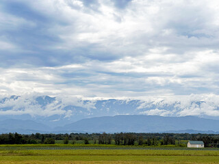 Fototapeta premium A breathtaking view showcases a vast green field bordered by mountains under a dramatic, cloudy sky. The peaceful rural setting includes a small white building in the distance.