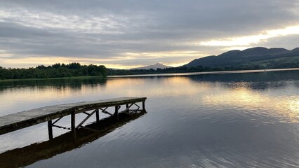 A tranquil sunset over Lake of Menteith in Scotland. A wooden dock stretches out into the calm water, reflecting the colorful sky and distant Ben A'an mountain - UK