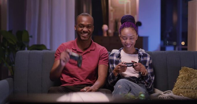 Casual happy love couple of young african woman and man holding gamepads wireless devices playing jostick console videogame on TV. Family portrait.