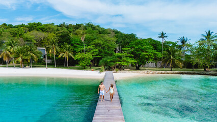 Couples stroll along a wooden pier leading to Koh Kham Thailand serene white sands, surrounded by emerald waters and lush tropical greenery. Perfect for a romantic getaway or peaceful retreat.
