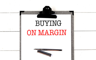 Clipboard with the words buying on margin and a silver pen on white wooden background 