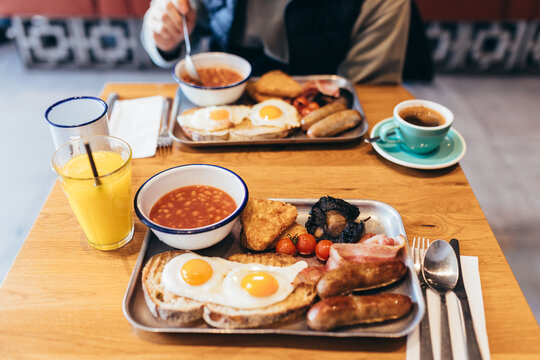 POV shot of a full English breakfast on metal trays: fried eggs, sausages, baked beans, hash browns, bacon, mushrooms, tomatoes, juice, and coffee. Soft focus