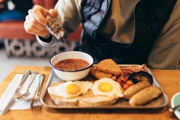 A man adding pepper to his traditional full English breakfast served on a metal tray, including...