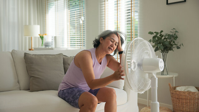 Asia adult people middle age old woman sit at sofa home feel hot flash fever use fan waving hand blowing wind face elder health care summer heat aging issue. Sad pain sweat tired stroke suffer stress.