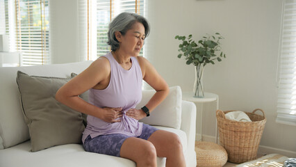 Asia adult middle age woman sit at sofa home feel pain elder gut health care aging system medical...