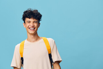 cheerful young man smiling with curly hair wearing a beige t shirt and yellow backpack against a bright blue background, capturing a sense of joy and positivity