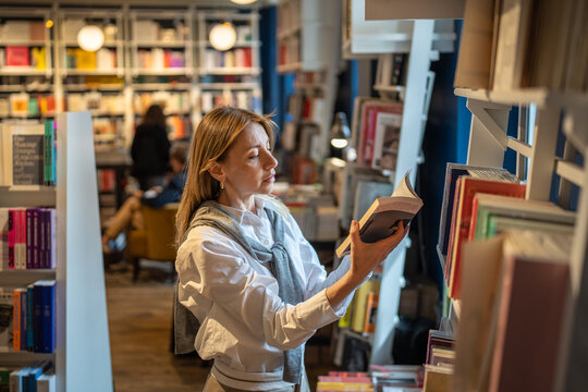 Thoughtful woman customer chooses and reads books from shelves in bookshop. Book worm female and reading hobby, browsing literature, book lover, looking for textbooks for study as adult student