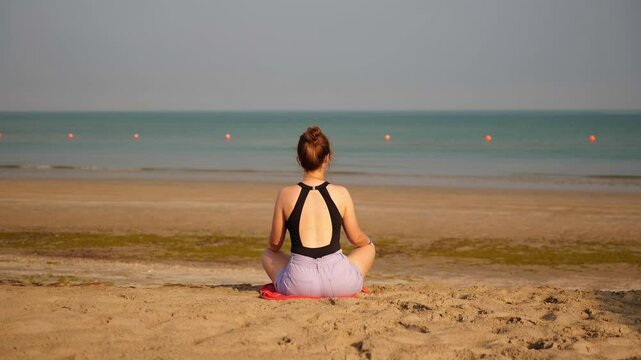 A young woman meditates on the beach, sitting in a calm yoga pose. The gentle waves and open sky create a harmonious and relaxing environment. Sitting on a towel with the ocean.