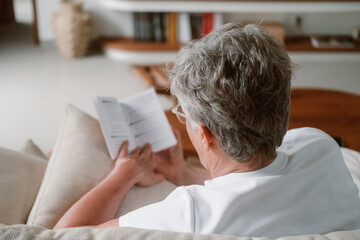A senior woman with white hair and glasses sits on a sofa in the living room and reads a paper...