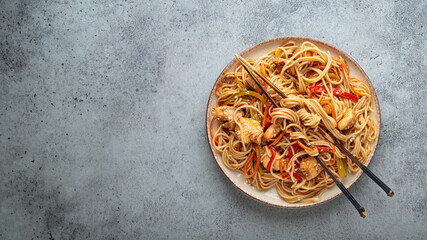 Udon noodles stir fried with chicken meat, carrots, bell peppers and other vegetables are served on a plate with chopsticks on a gray background and sprinkled with sesame seeds, copy space