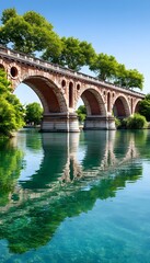 Fototapeta premium Red brick arched bridge over calm river, reflecting in clear water under sunny sky, lined with lush green trees