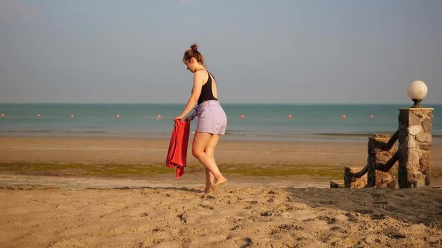 A young woman walks barefoot on the sandy beach, holding a towel. The calm sea and clear sky create a serene and peaceful atmosphere. A woman strolls along the sandy shore, carrying a towel.