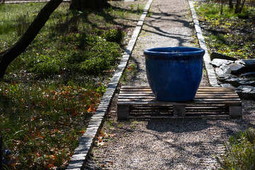 Fototapeta premium Large empty blue ceramic flowerpot placed on wooden pallet in landscaped park. Suitable for gardening blogs, design mockups, or landscaping product ads.