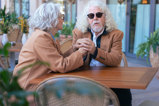 Caucasian senior couple holding hands at a wooden outdoor cafe table. They share an emotional moment, dressed in brown jackets. Concept of love, connection, maturity, and peaceful lifestyle.  - Powered by Adobe