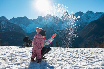 Mother and young daughter playfully toss sparkling snow powder into bright sunlight, enjoying...