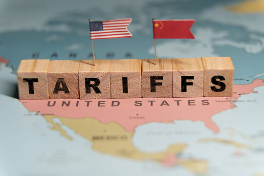  Wooden blocks spelling TARIFFS placed on a map of the United States with US and China flags, symbolizing trade tensions.