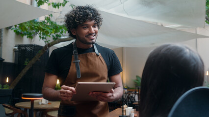Smiling Indian man Arabian latino guy friendly cafe worker barista in apron chef cafe customer service talk client order food lunch dinner in restaurant digital tablet serving visitor in coffee shop