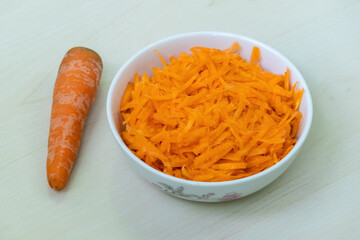 Grated carrot in a white bowl, on a light wooden surface, with a whole carrot placed beside.