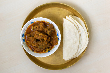 Tasty mutton curry and roti (flatbread) served on a brass plate on a light wooden surface – a very delicious Bengali meal. Top view.