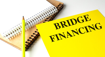 Yellow folder with the words bridge financing next to notebooks and pencil on white background 