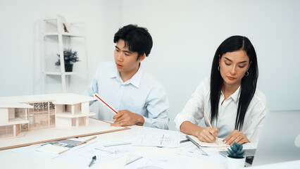 Professional male engineer measure house model by using ruler while beautiful cooperative coworker working together by writing on blueprint. Creative design and teamwork. Closeup. Immaculate.