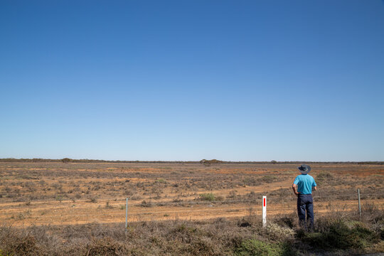 A man with hat looks out over an arid, harsh environment used as farmland with poor results owing to the harsh climate, under a blue sky near the city of Mildura in Victoria, Australia.