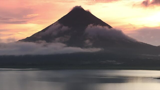 Witness a breathtaking aerial view of the stunning Arenal Volcano at sunrise, beautifully surrounded by mist, while the serene Laguna de Arenal glistens in the early light, located in Costa Rica