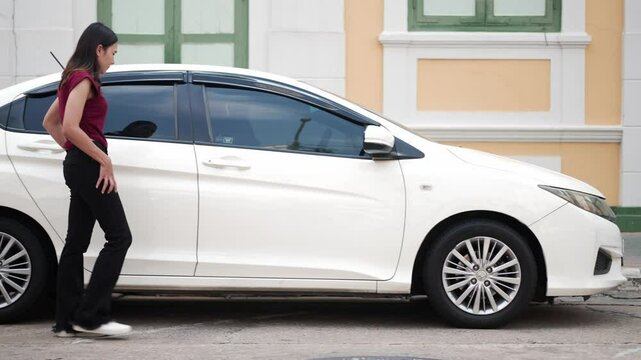 young asian woman walks beside car, checks pocket with confused expression, then spots key on the ground and bends down to pick it up, urban background