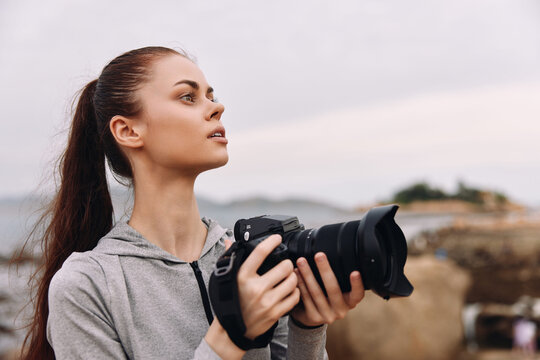 Young woman with long hair holds a camera, dressed in a gray hoodie, gazing thoughtfully at the horizon in a coastal setting with a moody sky