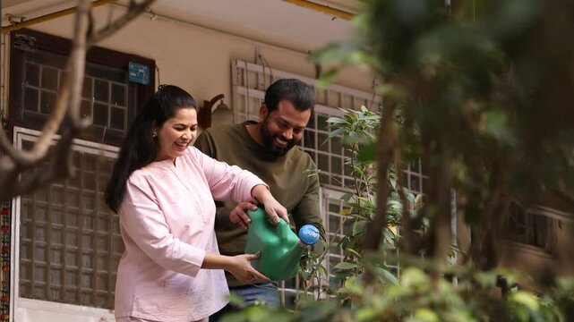 Young indian couple watering plants together outside their house garden in summer.Husband wife spend time together do Gardening hobby activity. Bonding and relationship. People of india. 4k