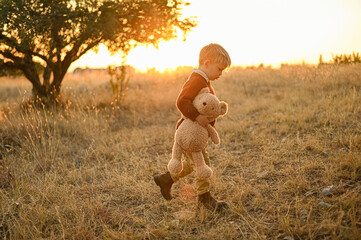 Little boy with a teddy bear walking through an olive grove at sunset. Warm rural scene with yellow grass, soft light, and peaceful childhood atmosphere.