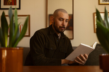 Fototapeta premium A thoughtful man with a beard sitting in a cozy, plant-filled room, reading a book. Warm, lifestyle atmosphere with a vintage aesthetic.