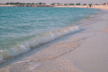Gentle waves softly wash the sandy shore of a tranquil UAE beach, inviting relaxation and leisure. Distant figures enjoy the serene ambiance of the coastline in a peaceful setting