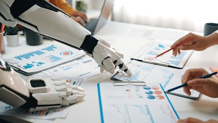 A robotic arm assists human colleagues in analyzing business data, highlighting the fusion of advanced technology and teamwork for effective decision-making in a modern workspace.Noogenesis