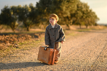 A boy in a coat and cap stands on a rural road with a suitcase, evoking a vintage feel of travel, nostalgia, and simple country life. A moment of quiet reflection and new beginnings.