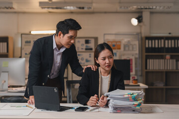 Young businessman comforting his stressed colleague sitting at office desk with a big pile of paperwork and looking worried, showing support and empathy in a challenging work environment