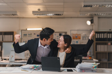 Celebrating success, two asian business colleagues are raising their arms in excitement while surrounded by paperwork and a laptop on the desk, embodying teamwork and achievement in the office